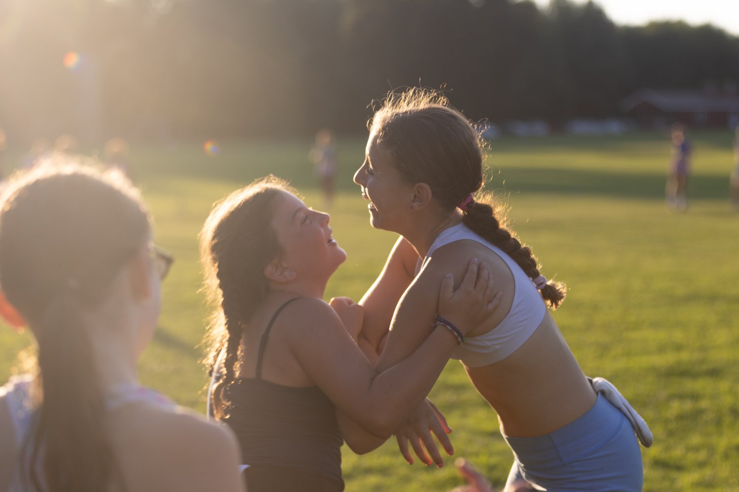 Two campers laughing face to face in backlit evening sun, a third figure framing the foreground