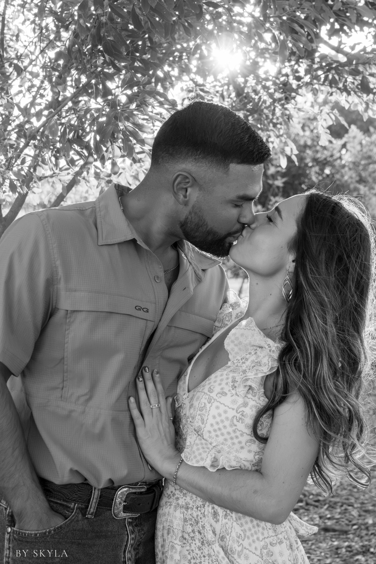 Black-and-white portrait of a couple kissing beneath sunlit trees, photographed by By Skyla