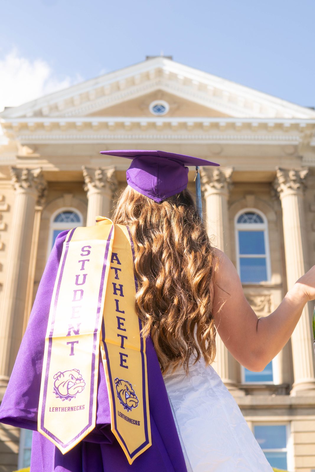 Graduate in cap and athlete stole with arms raised at campus columns