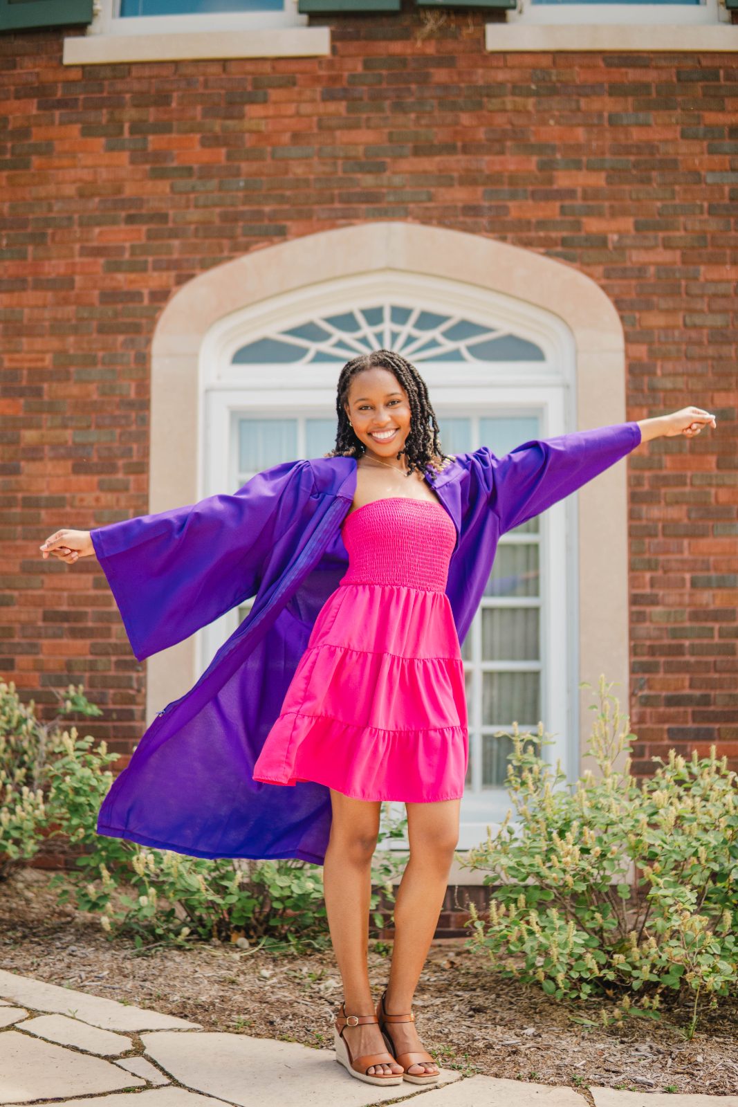 Graduate in a pink dress and purple gown with arms outstretched